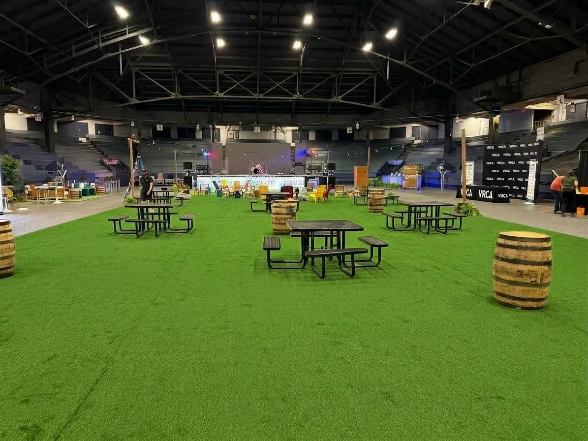 A wide field of green turf across an arena floor under a steel roof, set with black picnic tables and wooden barrels, the empty seating rising behind it