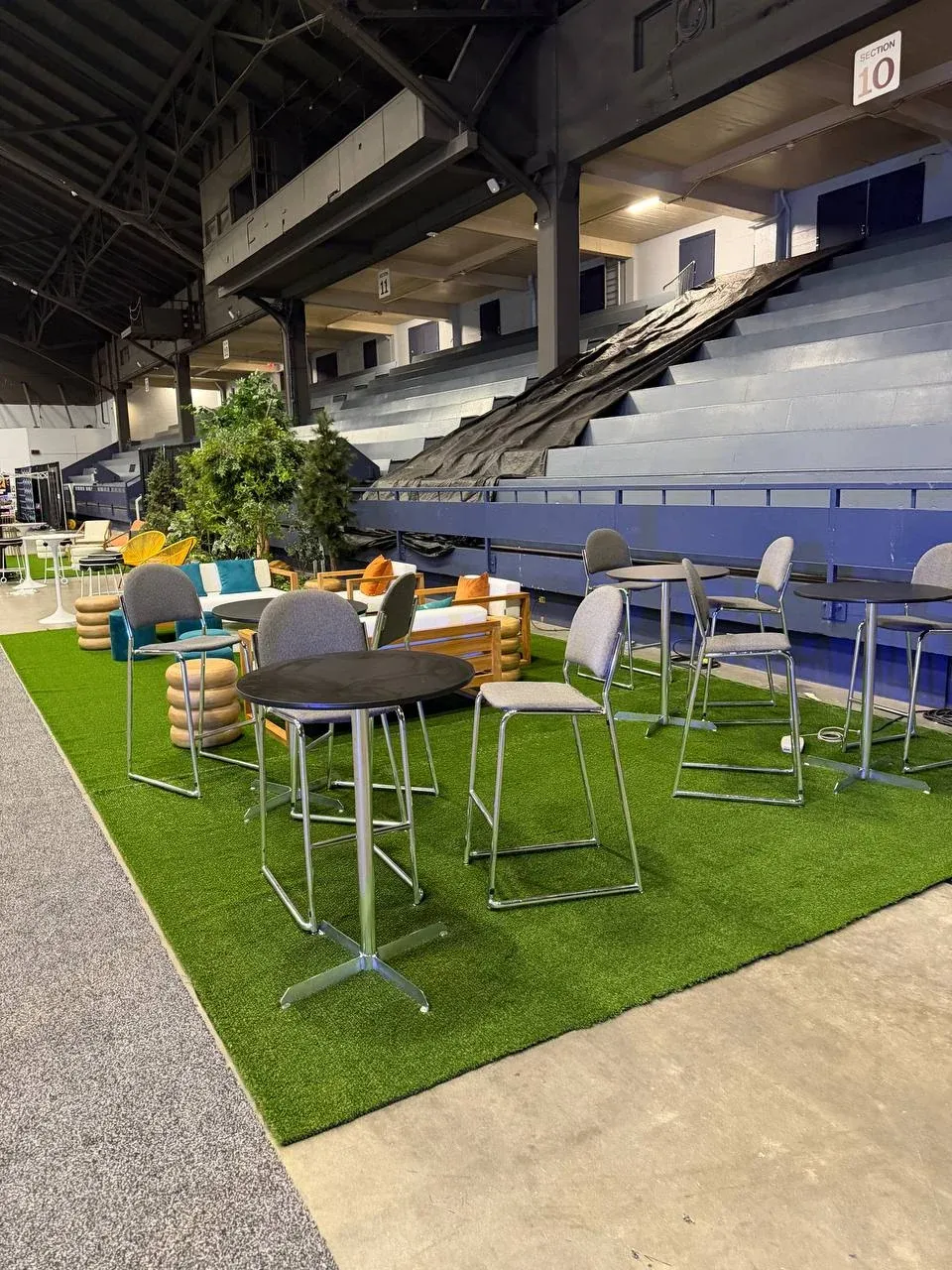 A turf lounge beside arena seating, chrome bar tables and grey upholstered stools on it and its straight edge running along a bare concrete aisle