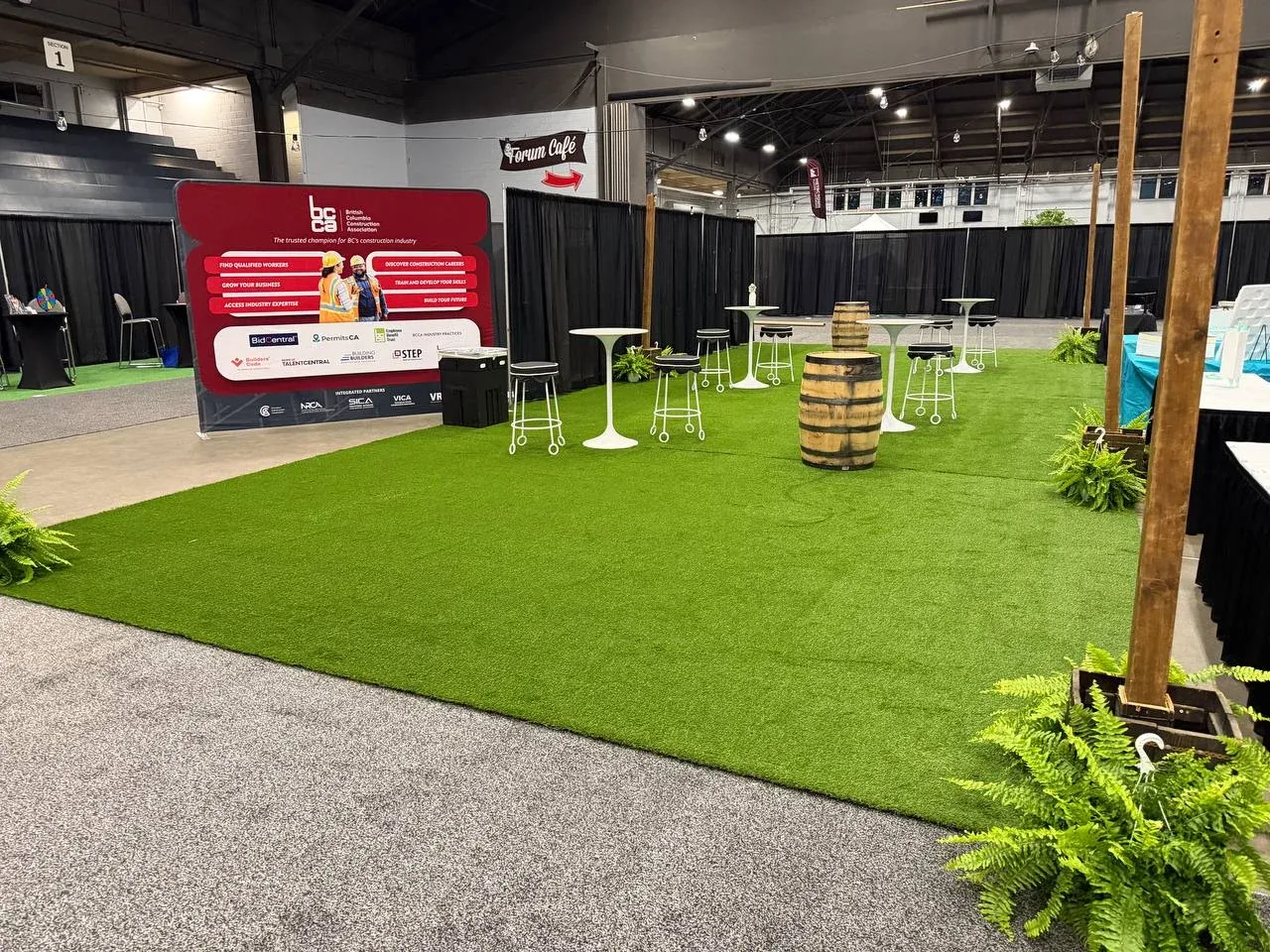 A rectangle of turf at a trade show with white bar tables, stools and a barrel on it, ferns along the edge where it meets grey carpet