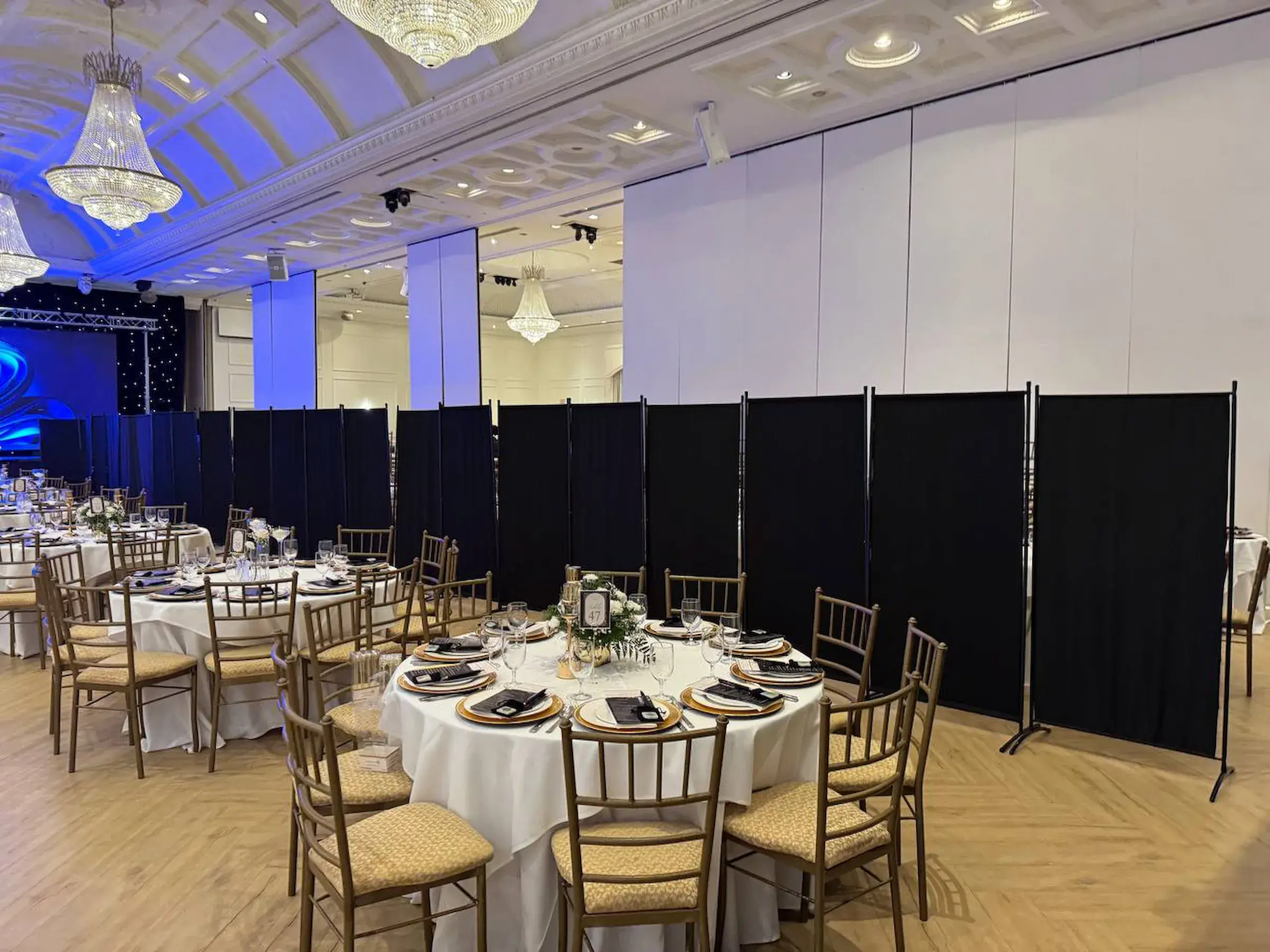A long run of black freestanding divider panels screening one side of a ballroom, with linened round tables laid for dinner and gilt banquet chairs in front of them under chandeliers