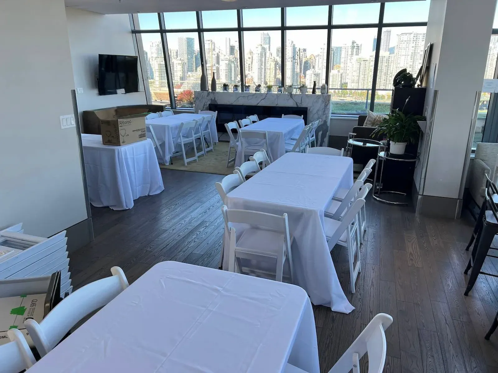 White folding chairs at rectangular tables under white linen in a room with floor-to-ceiling windows over a city skyline
