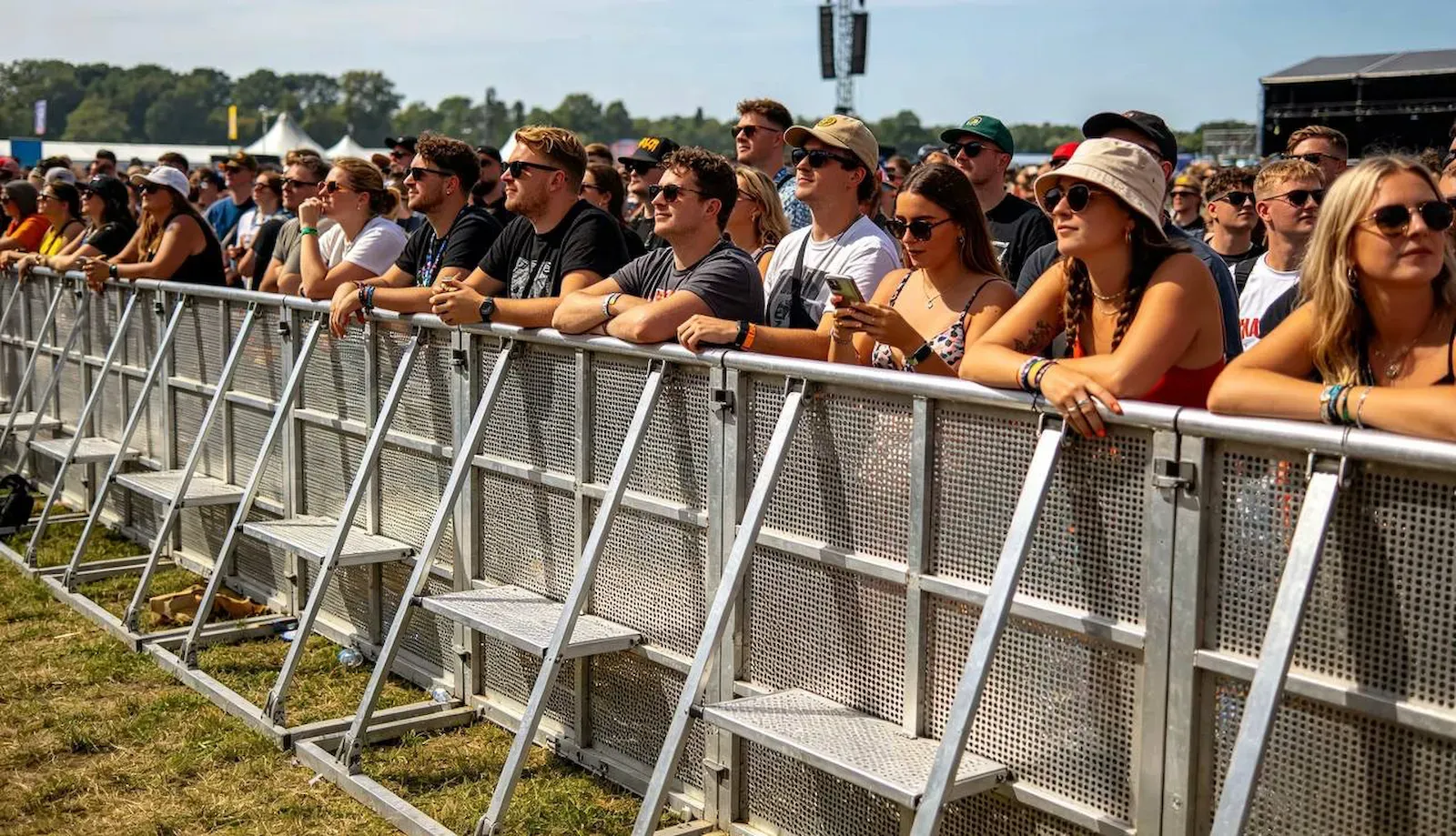 A line of aluminium stage barrier holding the front row at an outdoor show, people leaning on the top rail in the sun