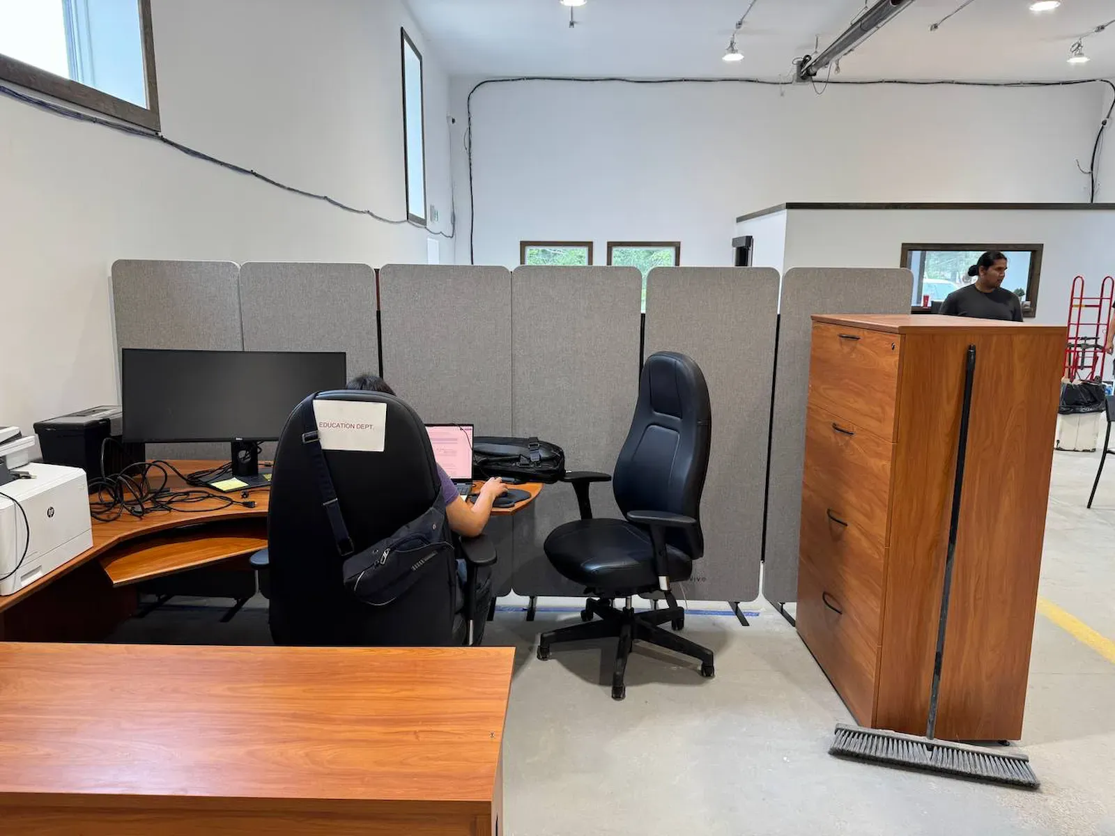 Grey acoustic panels standing as a screen along a run of office desks, a printer and a monitor on one side of them and a filing cabinet closing the end