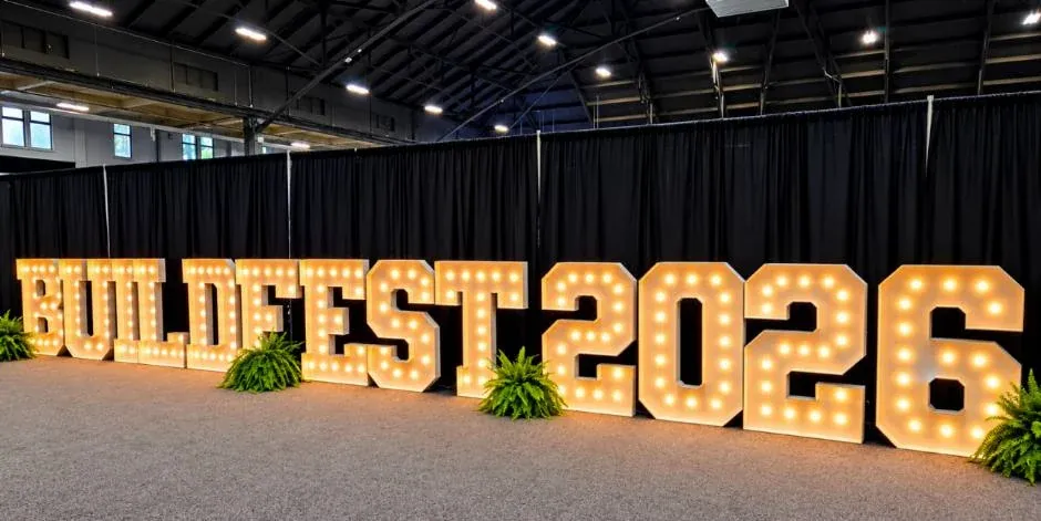 Four-foot marquee letters spelling a show name and the year in a row across a trade show hall, every bulb lit warm white against black drape, ferns set between them