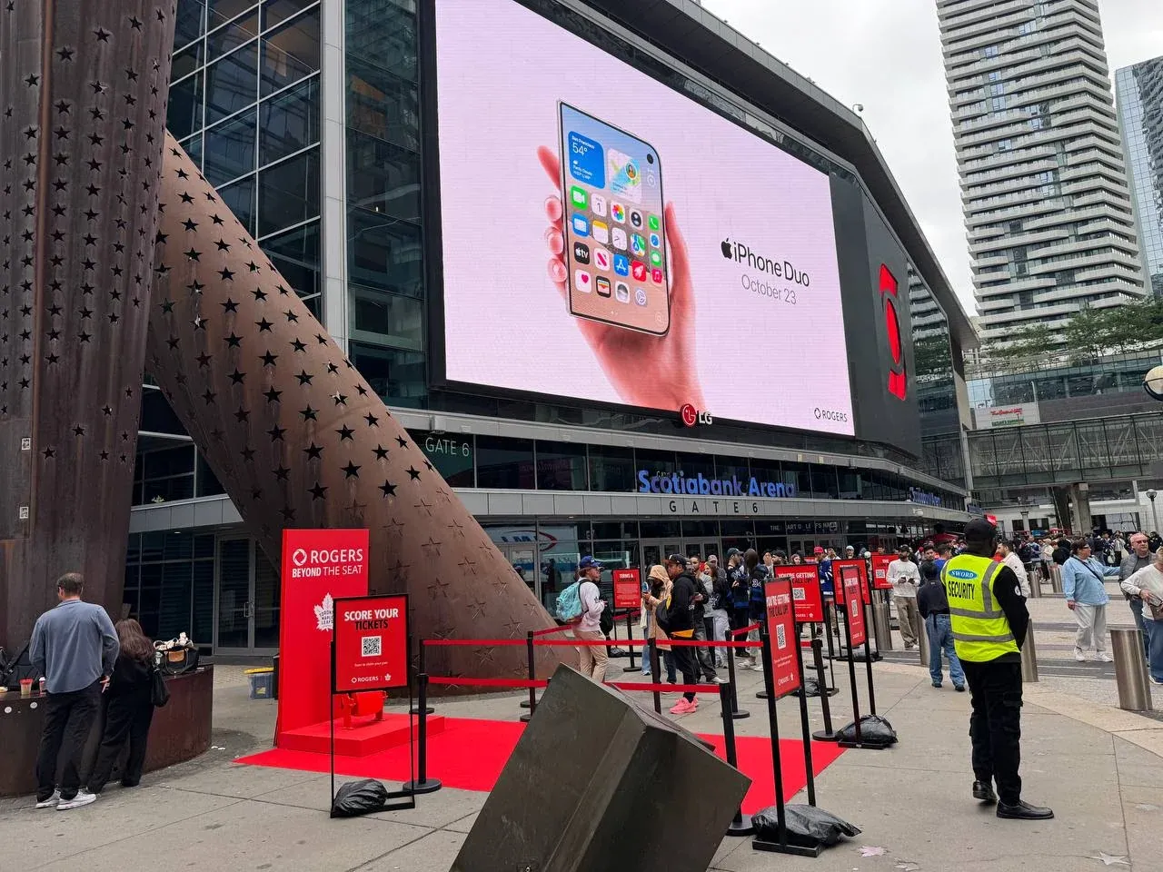 Black posts with red belts running a queue across a paved plaza outside an arena entrance, sandbags on the bases and red sign frames set along the line
