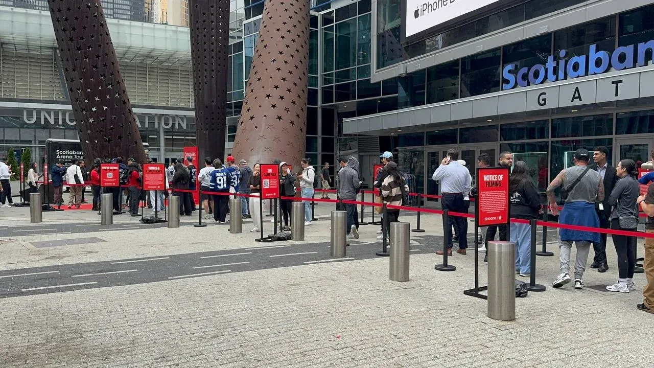 A long red-belt line on black posts stretching across a plaza towards an arena gate, people waiting along it between steel bollards