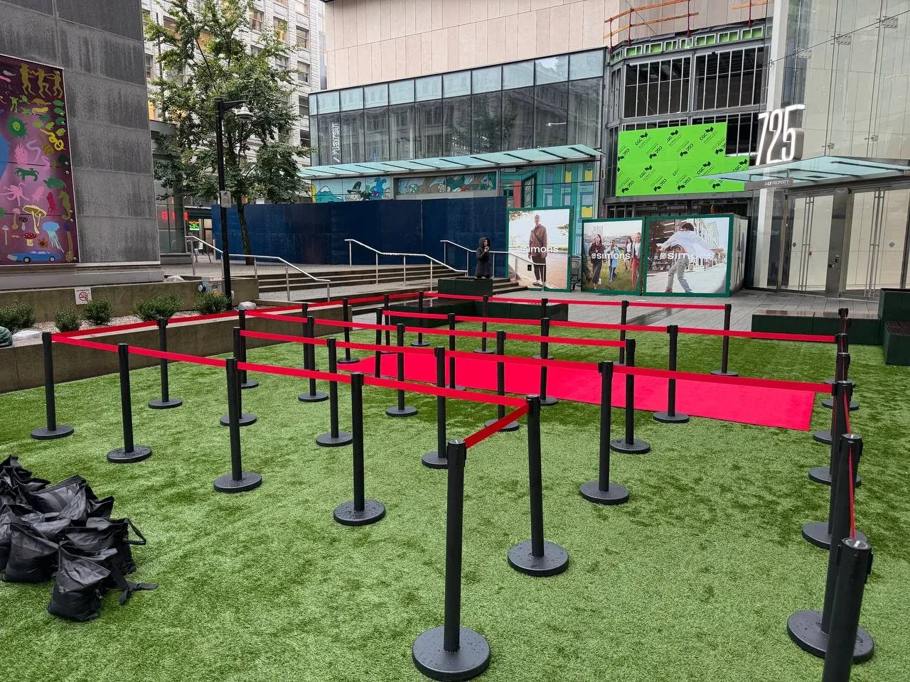 Black posts with red belts set out as a switchback queue on artificial turf in a city plaza, a red carpet runner down the middle and sandbags piled beside it