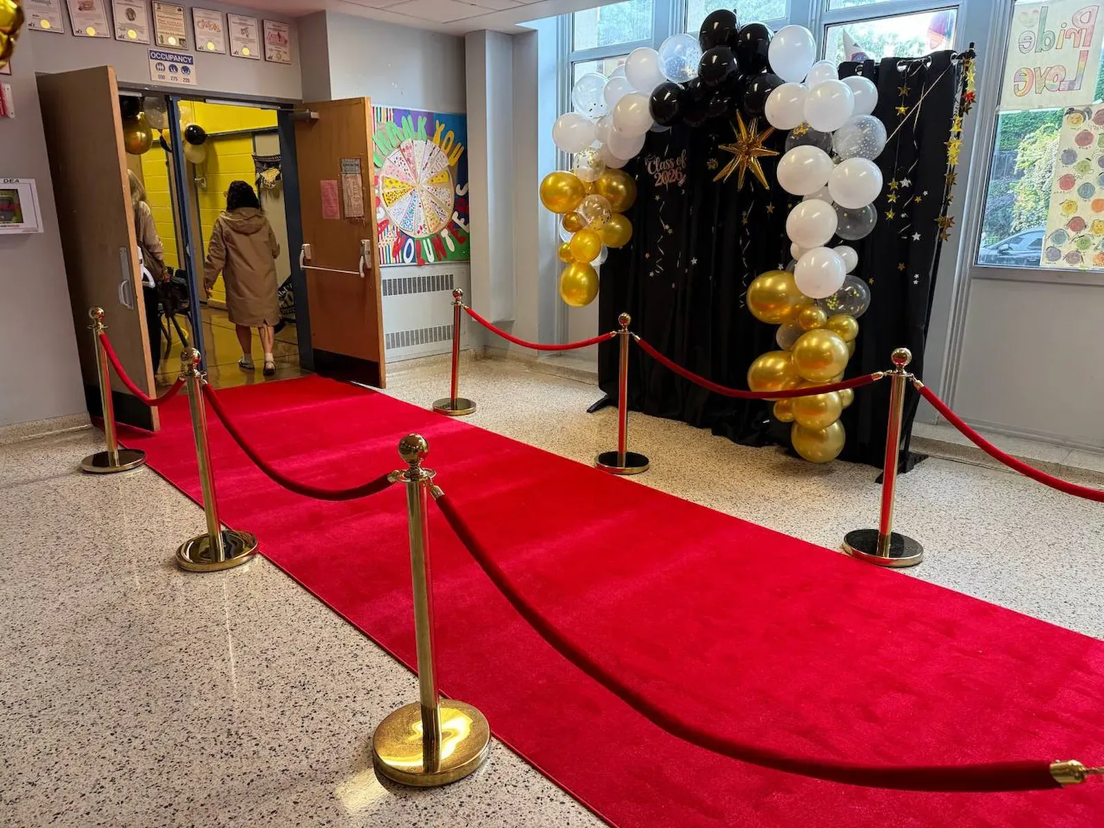 A red runner laid the length of a school corridor between gold posts and red ropes, a balloon arch at the far end