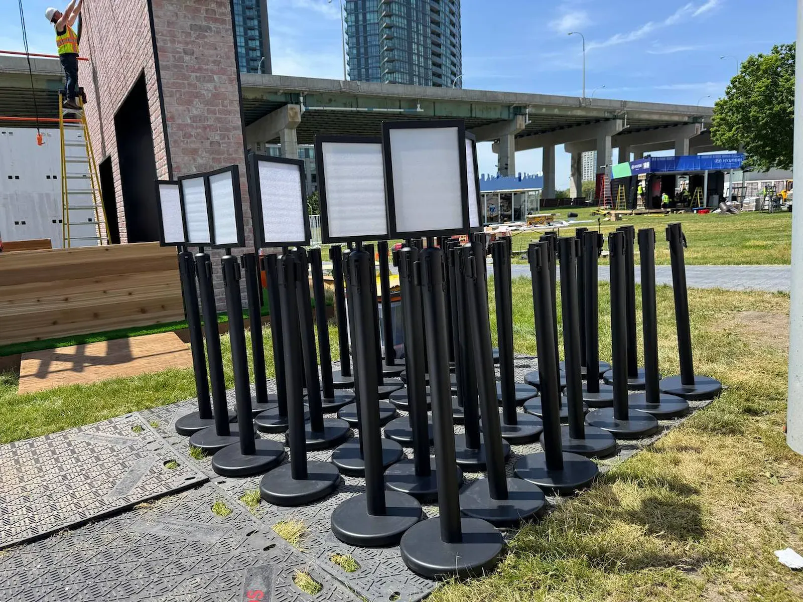 Dozens of black belt posts standing on ground-protection matting during a build, several of them carrying printed sign frames at head height
